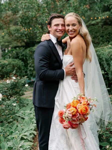 Couple portrait of bride and groom hugging and smiling, bride in strapless dress with veil holding bouquet with ribbon in garden lights