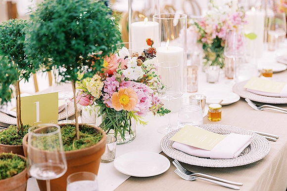 Reception tablescape with wedding table setting, colorful floral centerpiece in glass vase, taper candles, woven chargers, and table number on neutral linen