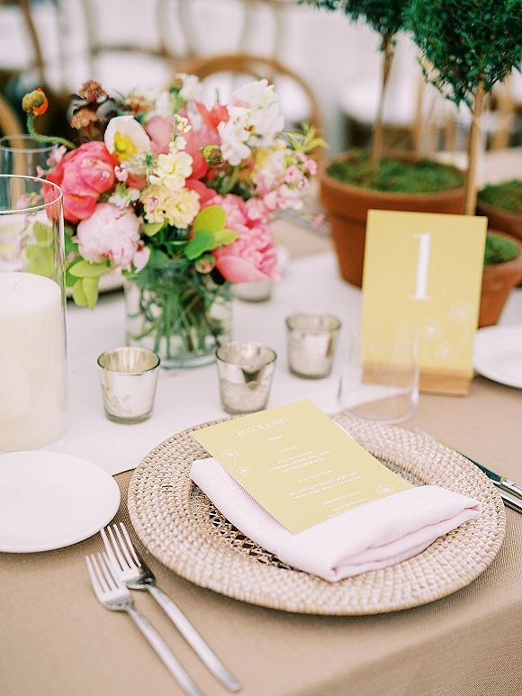 Reception tablescape with wedding place setting featuring a floral centerpiece in glass vase, menu and table number cards, and candlelight on neutral linens