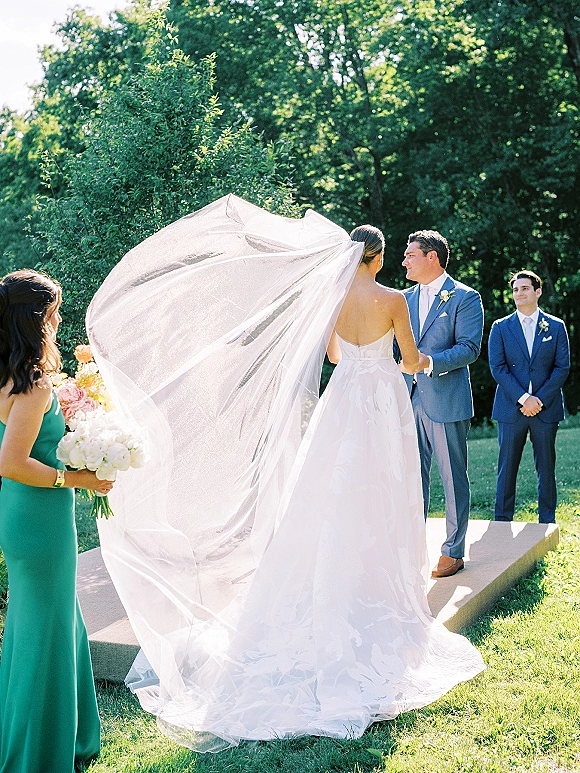 Wedding vows as bride and groom hold hands on a sunny outdoor wedding ceremony platform, her veil blowing beside bridesmaids with bouquets