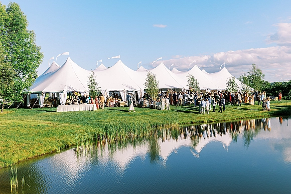 Wedding reception tent with sailcloth wedding tent flags, banquet tables and lounge seating set on a lawn by a pond under blue sky