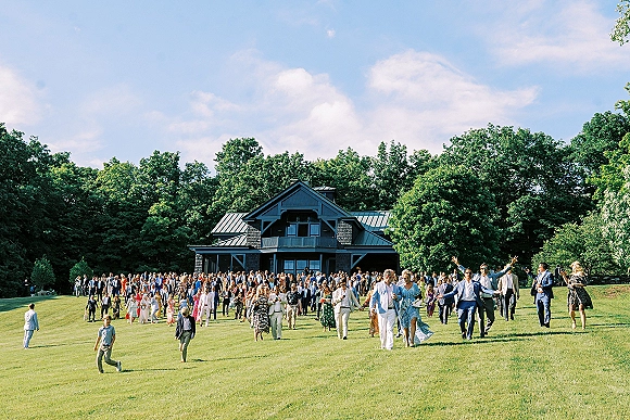 Wedding guest group photo of a large crowd in suits and colorful dresses, some with sunglasses, gathered on a grassy lawn by a house under blue sky