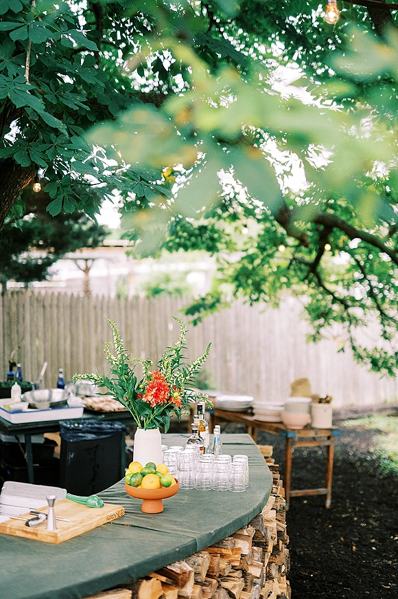 Outdoor bar setup with a green tablecloth and citrus bowl, glassware and liquor bottles under string lights in a fenced backyard