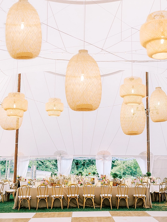 Reception tablescape with a long banquet table wedding setup, floral centerpieces and taper candles under woven pendant lanterns in a white tent