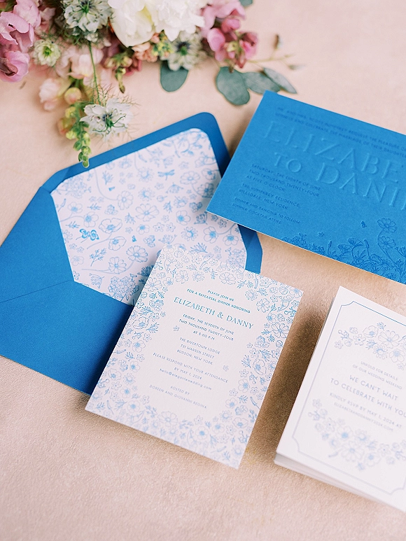 Wedding invitation suite with blue wedding invitations, embossed card and floral-lined envelope arranged with flowers on a linen tablecloth
