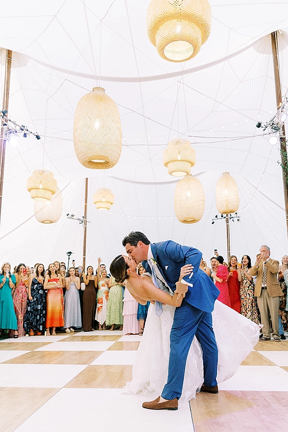 First dance kiss as the groom in a blue suit dips the bride in a strapless dress under string lights and hanging wicker lanterns in a tent reception