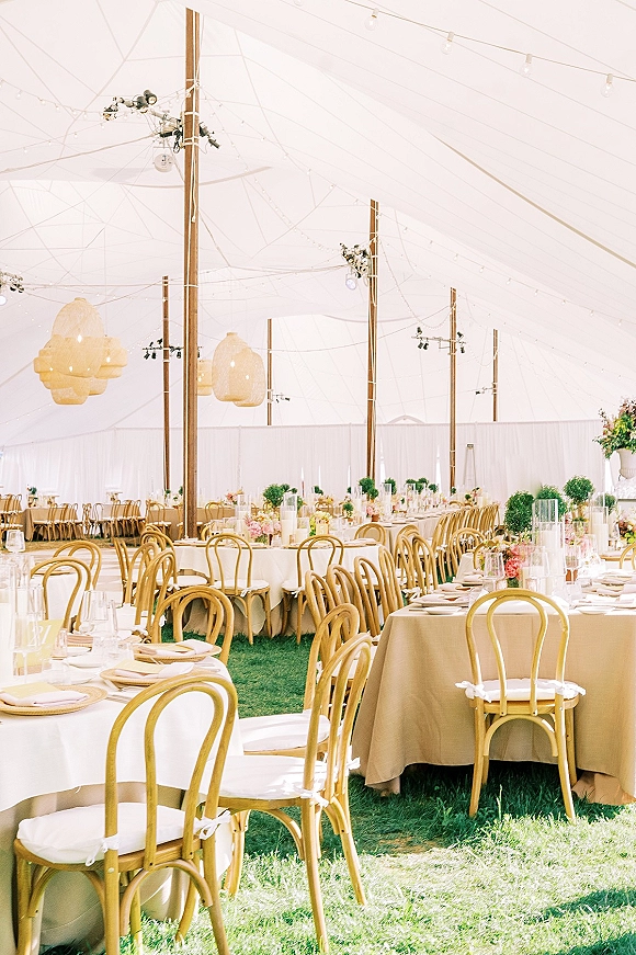 Reception tablescape with neutral linens on round tables, candles and floral centerpieces beneath string lights in a white tent on grass lawn
