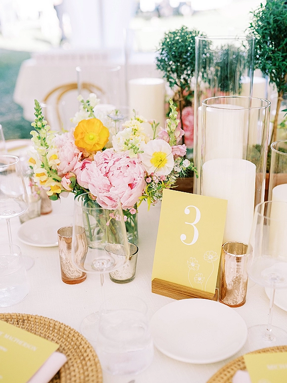 Reception tablescape with wedding table centerpiece of pink and yellow flowers in a glass vase, pillar candles, rattan chargers under a draped tent lawn setting