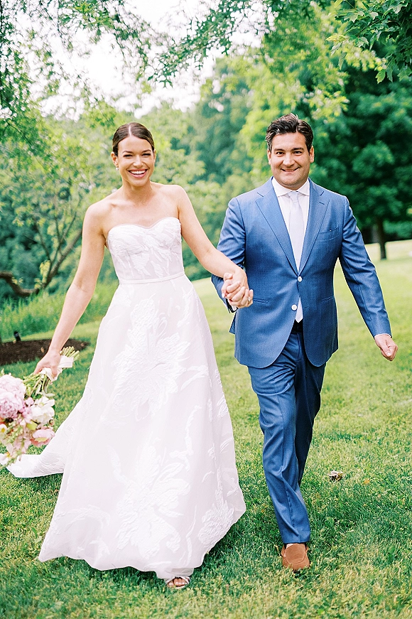 Couple portrait of bride and groom holding hands, bride in strapless dress with bouquet and groom in blue suit on a garden lawn