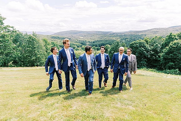 Groomsmen portrait with blue suits and one gray suit, walking together on a grassy field with mountains and cloudy sky behind