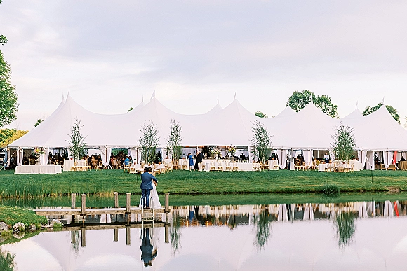 Couple portrait of bride and groom on dock, wedding dress and suit by a pond with water reflections beside a sailcloth tent reception setup