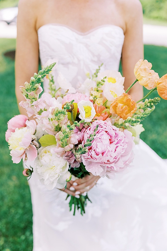 Bridal bouquet of pastel wedding bouquet blooms in pink and peach, with peonies and greenery held against a strapless lace dress on a green lawn