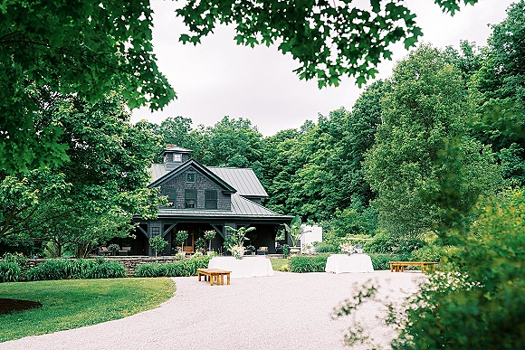 Outdoor reception setup with backyard wedding reception tables in white tablecloths, floral centerpieces, and benches by a cottage driveway and trees