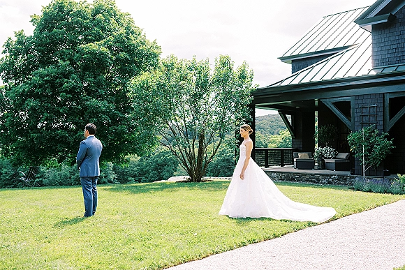 Wedding first look as bride approaches groom from behind, strapless gown with long train on lawn by modern cabin with mountains beyond