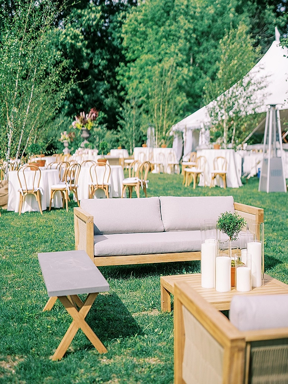 Outdoor lounge seating with a wood-frame sofa and cushions around a low table, candle accents, and a tented lawn reception setup
