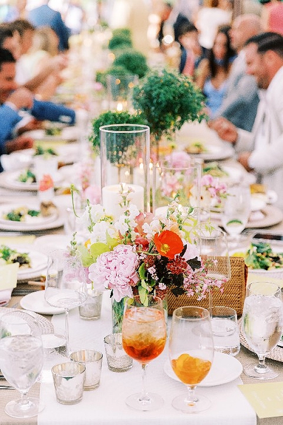 Reception tablescape with a long banquet table wedding setup, floral centerpieces and candlelit glass cylinders as guests dine indoors