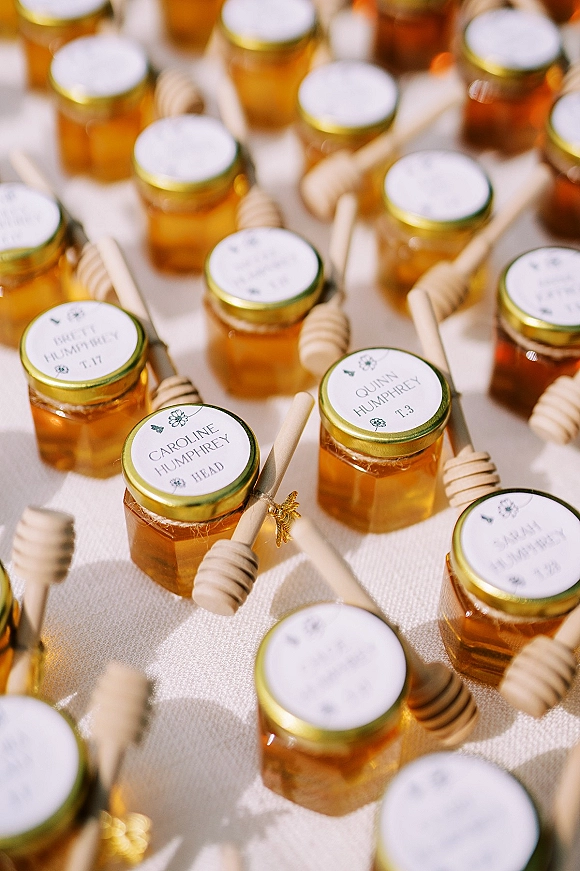 Wedding favors of mini honey jar wedding favors with gold lids, honey dippers, twine, and personalized round labels on white linen tablecloth