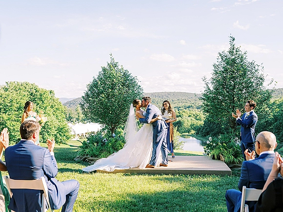 Ceremony kiss at an outdoor wedding ceremony as the bride and groom kiss on a platform, veil flowing with lake and hills behind
