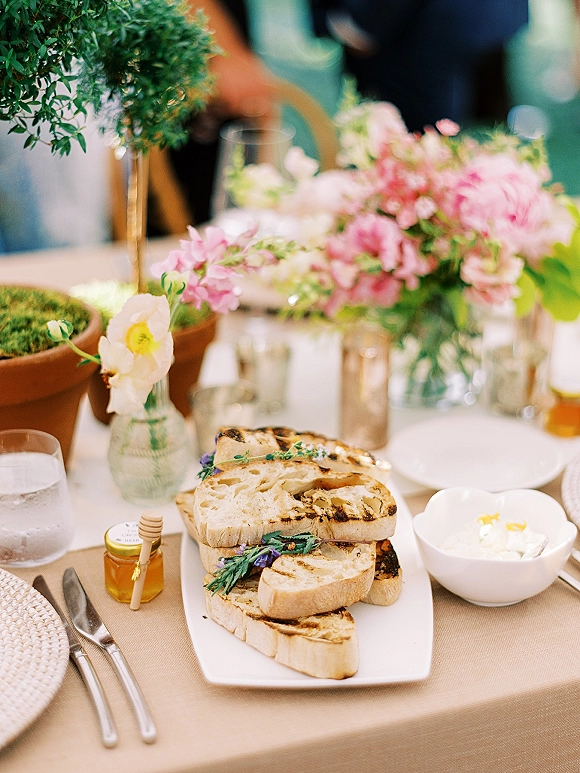 Reception tablescape with a wedding table centerpiece of pink florals and greenery, grilled bread platter, honey jar and dipper on beige linens