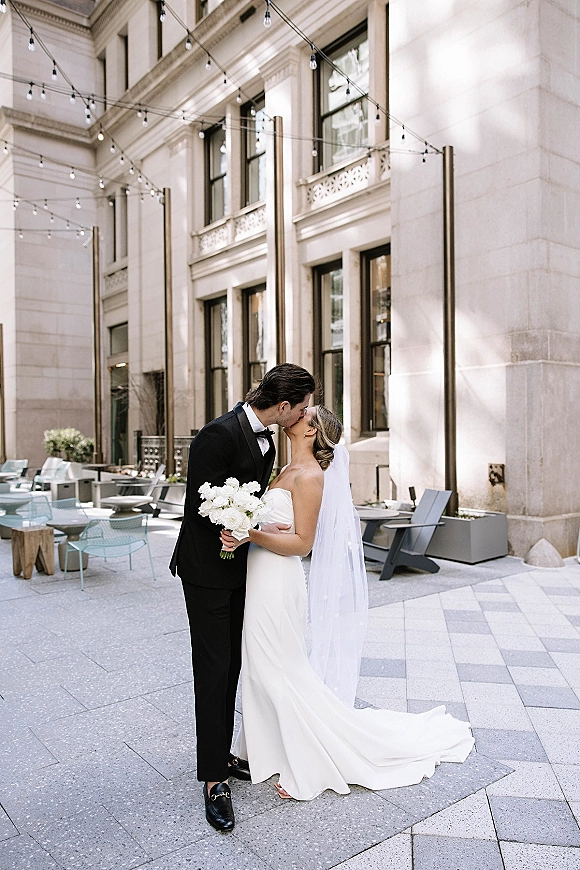 Wedding kiss portrait of bride and groom kissing, her long veil and white bouquet visible under string lights in a stone courtyard patio
