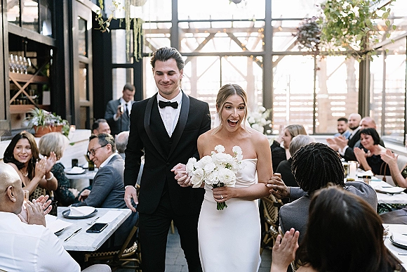 Wedding reception entrance as bride in strapless dress holds bouquet, walking hand in hand with groom in tuxedo past cheering guests in sunlit venue