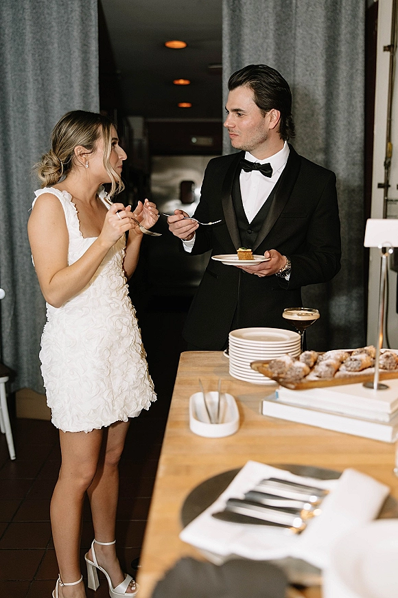 Reception dessert moment as bride in a short wedding dress and groom in tuxedo share cake bites at a wooden dessert table in hallway