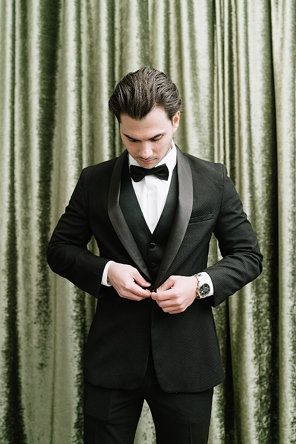 Groom portrait in a black tie groom tuxedo with satin shawl lapel, bow tie, waistcoat and wristwatch before a green velvet curtain backdrop