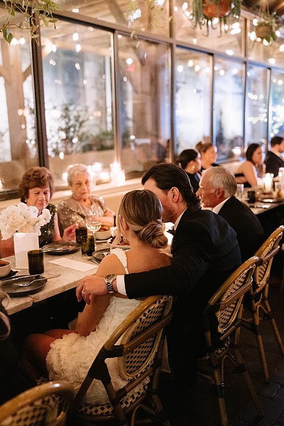 Reception moment as bride and groom at dinner seated at a long banquet table under string lights, with candles and floral centerpiece by glass windows