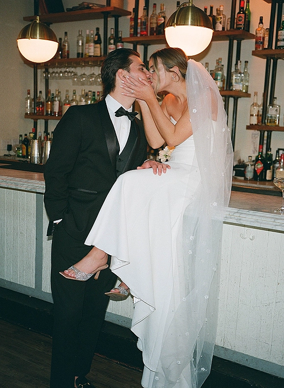Wedding kiss portrait of bride and groom kiss at an indoor bar, her long veil and bouquet visible beside liquor bottles and pendant lights