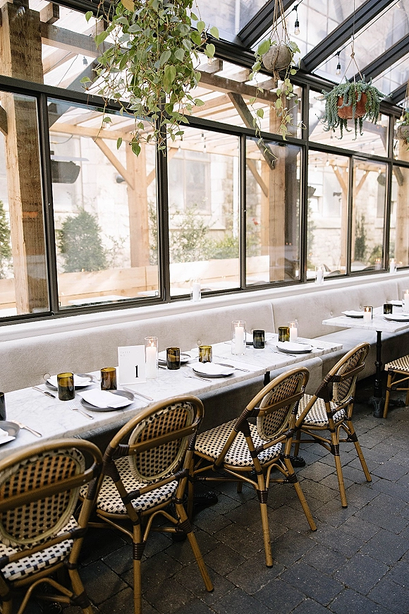 Reception tablescape with marble wedding table, white plates, amber tumblers, taper candles and greenery under a glasshouse ceiling with string lights