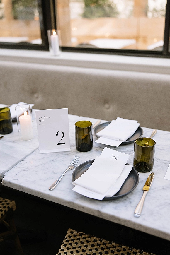 Reception tablescape on a marble wedding table with black plates, green tumblers, pillar candles, and menus by a daylight window banquette