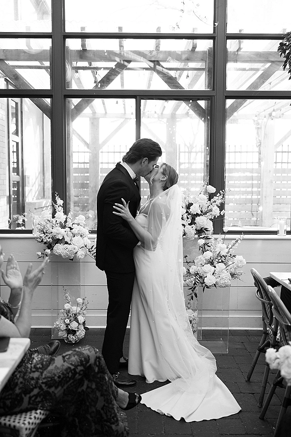 Wedding kiss as bride in strapless dress and veil kisses tuxedoed groom amid white roses on acrylic stands and aisle candles by grid windows