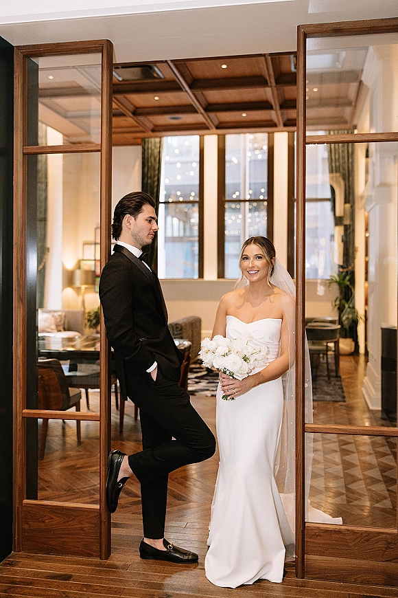 Couple portrait of bride holding a white rose bouquet and groom in black tuxedo, posed by wooden glass doors in a hotel lobby.