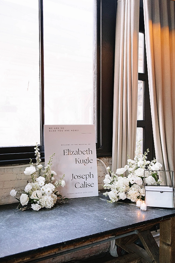 Wedding welcome sign with florals on a wooden table, accented by white roses, pillar candles, and a glass card box by large windows