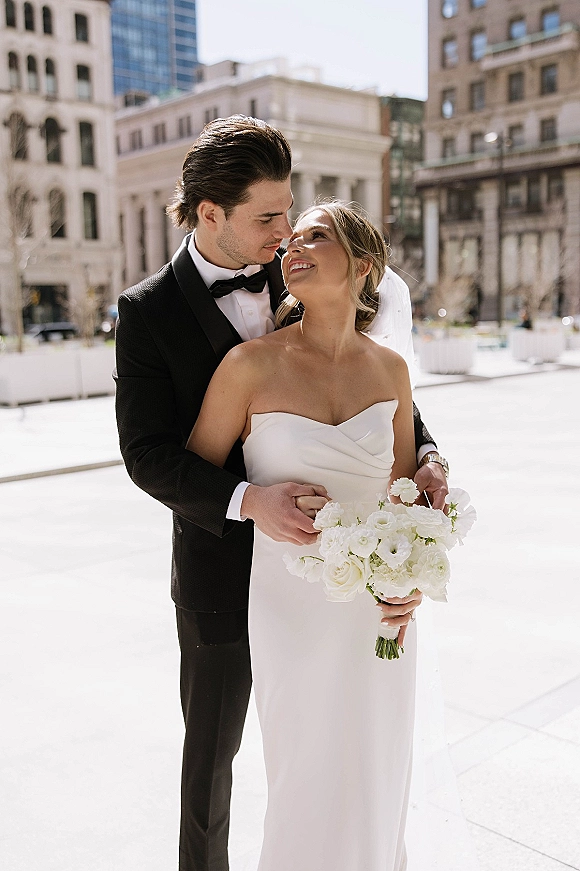 Couple portrait of bride and groom embrace, groom hugging from behind as she holds white rose bouquet on a bright city street with veil