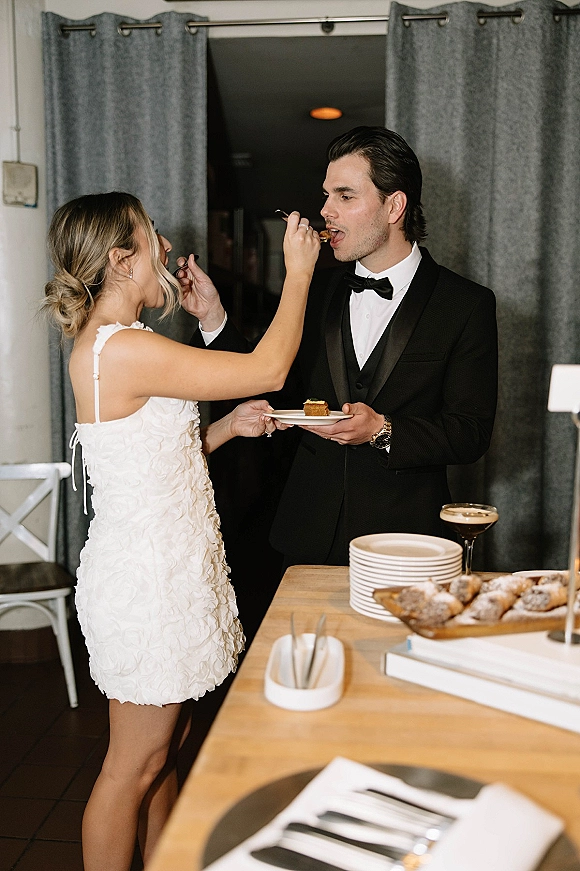 Cake cutting moment as bride in a short wedding dress feeds groom in a black tuxedo at a dessert table with gray curtains behind