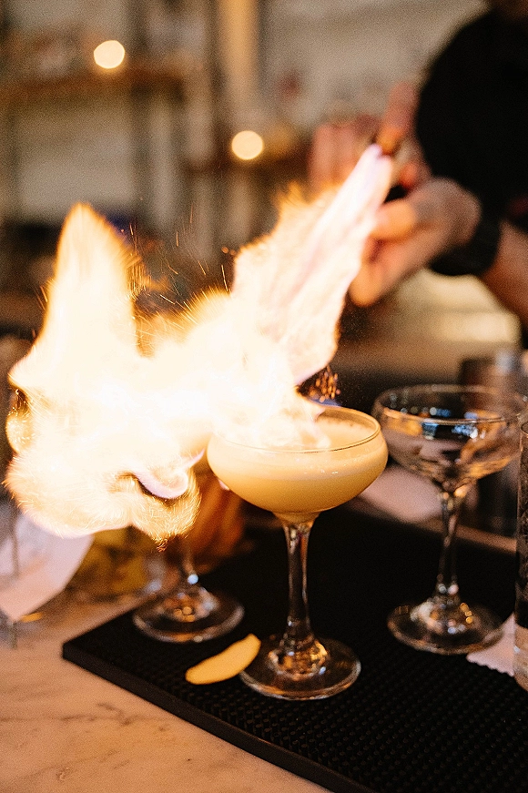 Flaming cocktail in a coupe glass with foam and open flame on a bar mat, bartender behind the counter under warm bistro lights