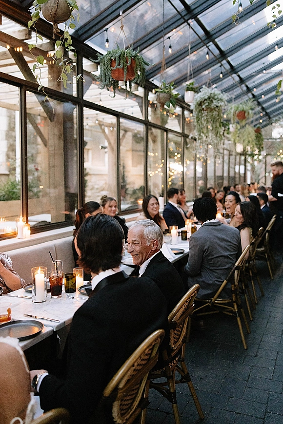 Wedding reception dinner with a long banquet table under string lights, candlelit place settings, and hanging plants in a glass greenhouse patio setting