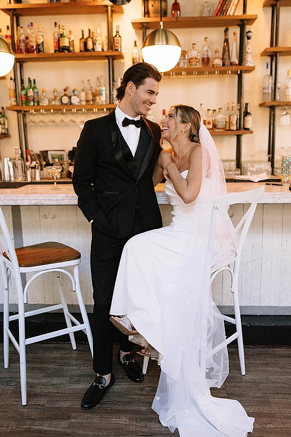 Couple portrait of bride and groom at bar, laughing as she sits on a bar stool in a strapless gown and veil by shelves of glassware