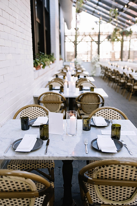 Reception tablescape on a marble wedding table with gray plates, white napkins, menu and place cards, and taper candles in glass hurricanes under string lights