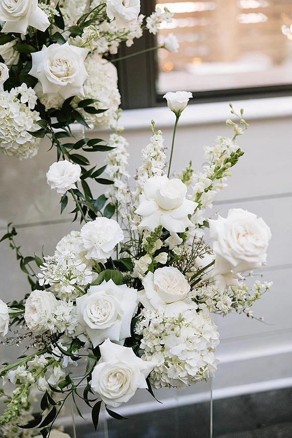 Wedding floral arrangement of white roses and hydrangeas with lush greenery against a white paneled wall and window in warm bokeh light