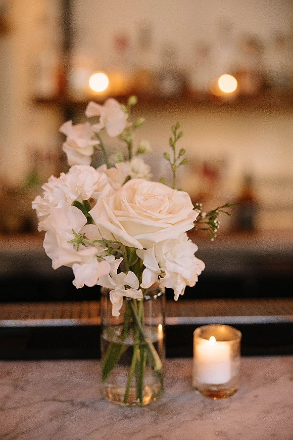 Wedding centerpiece with white roses and greenery in a clear glass vase beside votive candles on a marble bar top under warm lights