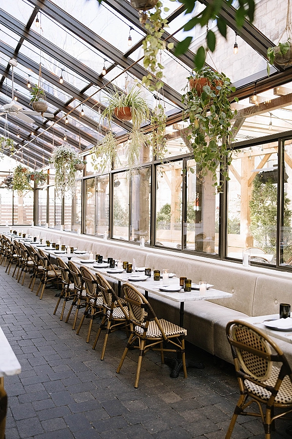 Reception tablescape with long banquet table setup, white plates and black napkins, votive candles, hanging plants and string lights in a glass greenhouse venue