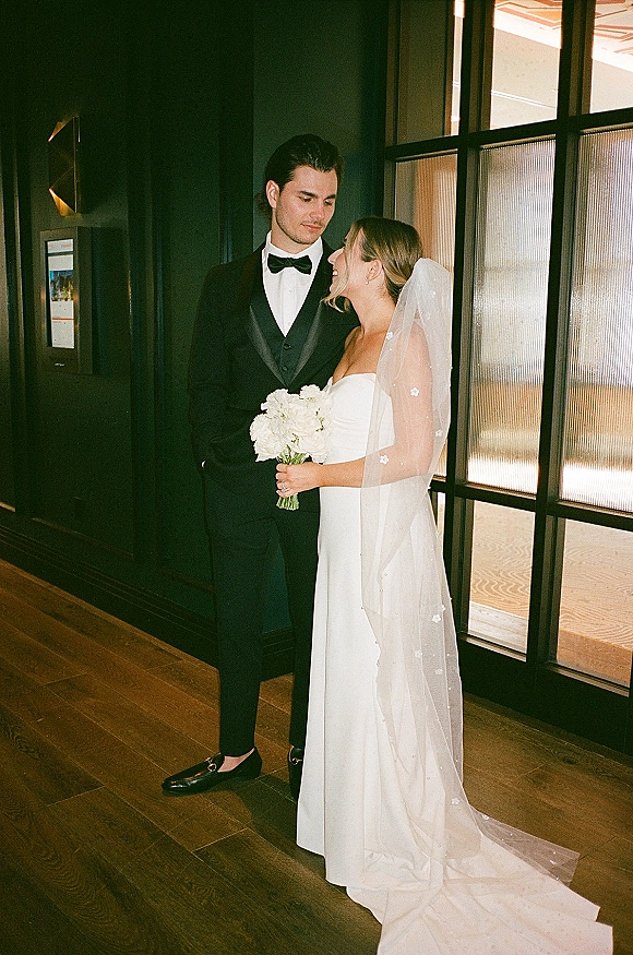 Couple portrait of bride looking at groom, holding a white bouquet in strapless gown and veil beside tuxedoed groom by a dark paneled wall