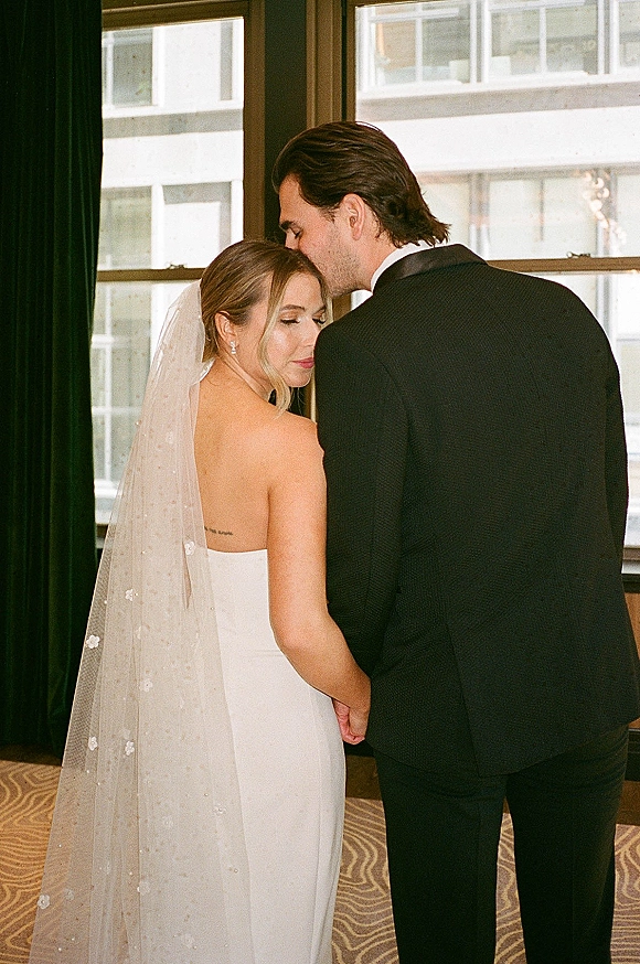 Couple portrait of bride and groom holding hands as he kisses her forehead, her long veil trailing by a large window with city view