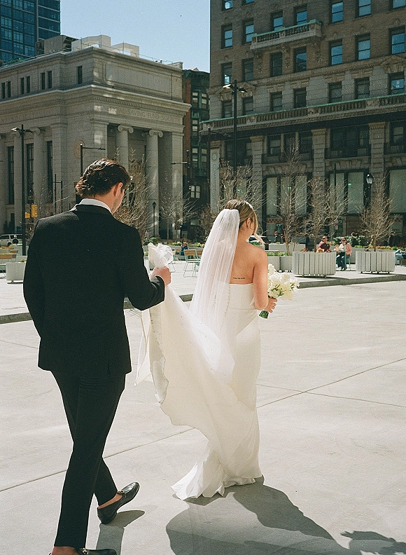 Wedding couple portrait of bride and groom walking in a city plaza, groom lifting her dress train, bouquet and long veil flowing under clear sky
