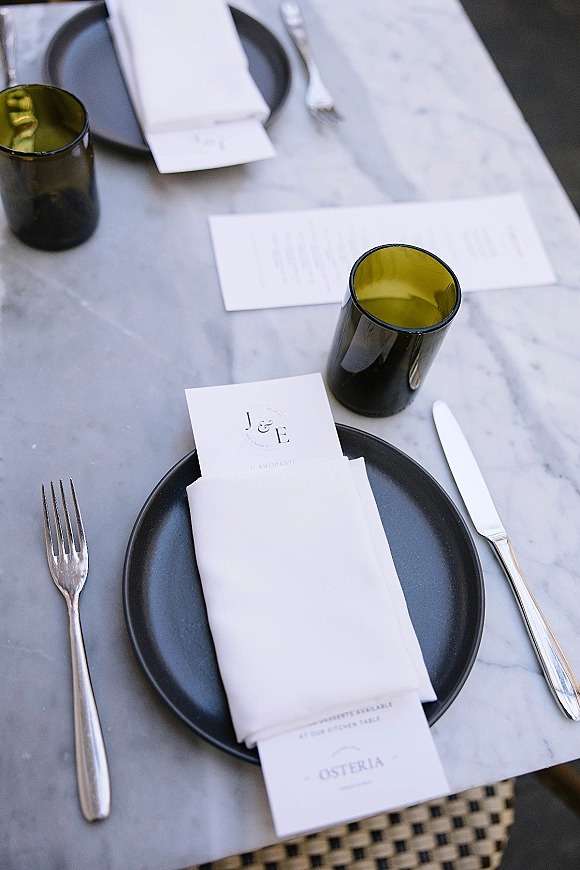 Wedding tablescape with black plates and crisp white napkins, menu and place cards, silver flatware and green glass tumblers on marble tabletop
