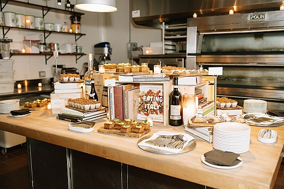 Dessert table styled as a wedding dessert table with assorted pastries and cake slices, candles, and stacked books in a bakery kitchen