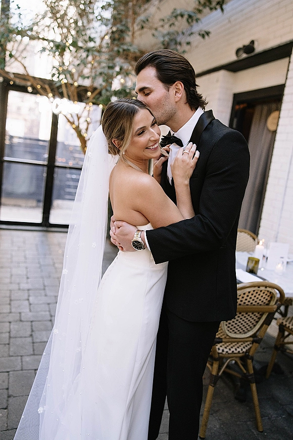 Couple portrait of groom kissing bride’s forehead as they embrace on an outdoor patio with string lights, her veil draped over strapless dress