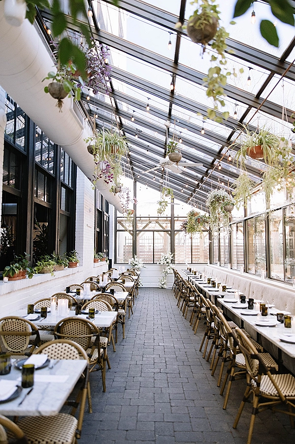 Reception tablescape in a greenhouse wedding reception with long white-linen banquet tables, rattan chairs, green glassware, taper candles, and hanging florals under string lights